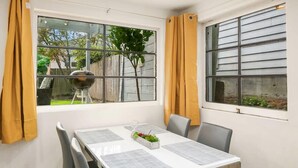 Comedor con una mesa lista para dos, una ventana con cortinas amarillas y vista a un patio trasero con una parrilla.