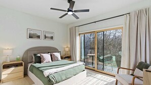 A bedroom with a bed, nightstands, a ceiling fan, a large window with curtains, and a balcony with chairs. - Overlook at Black Apple | On NWA Trail | Downtown (Bentonville)