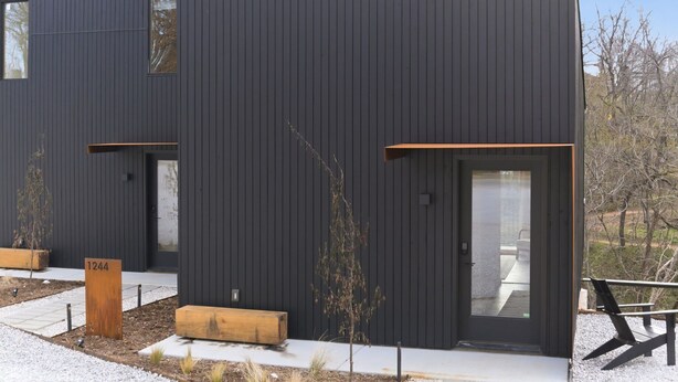 A modern building with a dark facade, a wooden bench, and a small tree in the foreground. - Overlook at Black Apple | On NWA Trail | Downtown (Bentonville)