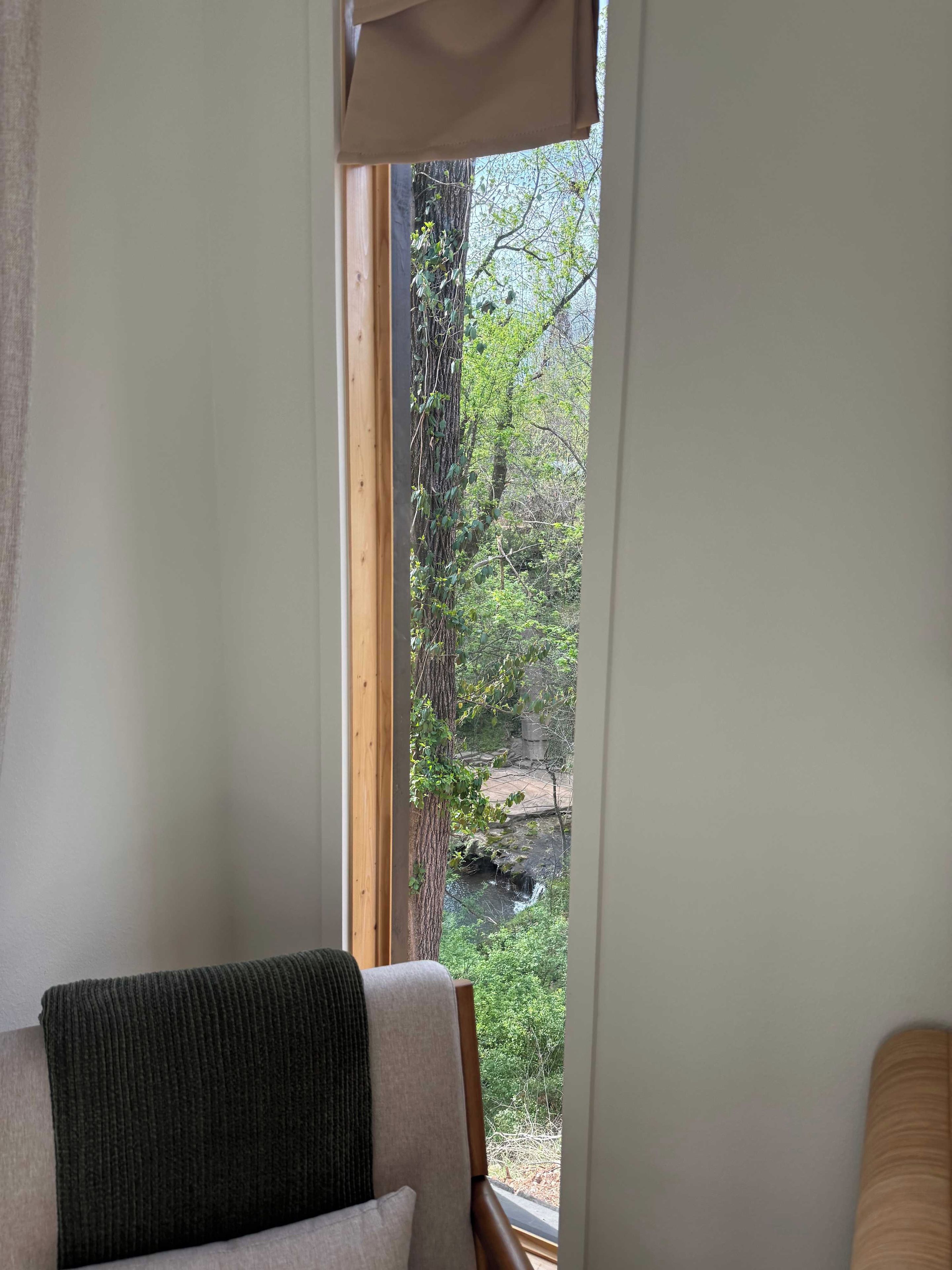 Una habitación con ventana que da a un paisaje natural, con árboles y un pequeño arroyo.