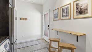 A hallway with a refrigerator, a door, a rug, a bench, and framed pictures on the wall. - Overlook at Black Apple | On NWA Trail | Downtown (Bentonville)
