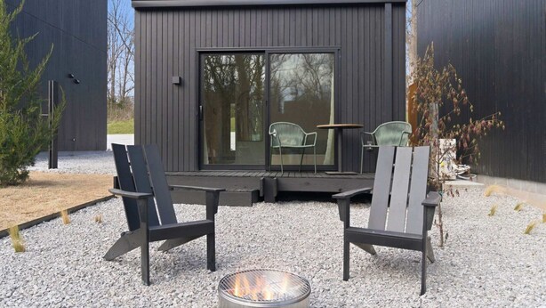 A modern outdoor seating area with two Adirondack chairs, a fire pit, and a glass door entrance to a building. - Vista at Black Apple | On NWA Trail | Downtown (Bentonville)