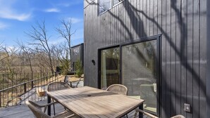 Eine Terrasse mit Holztisch und Korbstühlen, eine Glasschiebetür und Blick auf Bäume.