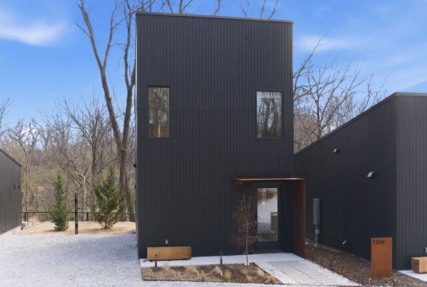 A modern building with a dark exterior, a small entryway, and a gravel driveway. - Trailhead at Black Apple | On NWA Trail | Downtown (Bentonville)