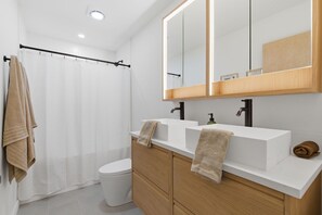 A modern bathroom with a white toilet, a shower curtain, a double sink vanity, and a large mirror. - Trailhead at Black Apple | On NWA Trail | Downtown (Bentonville)
