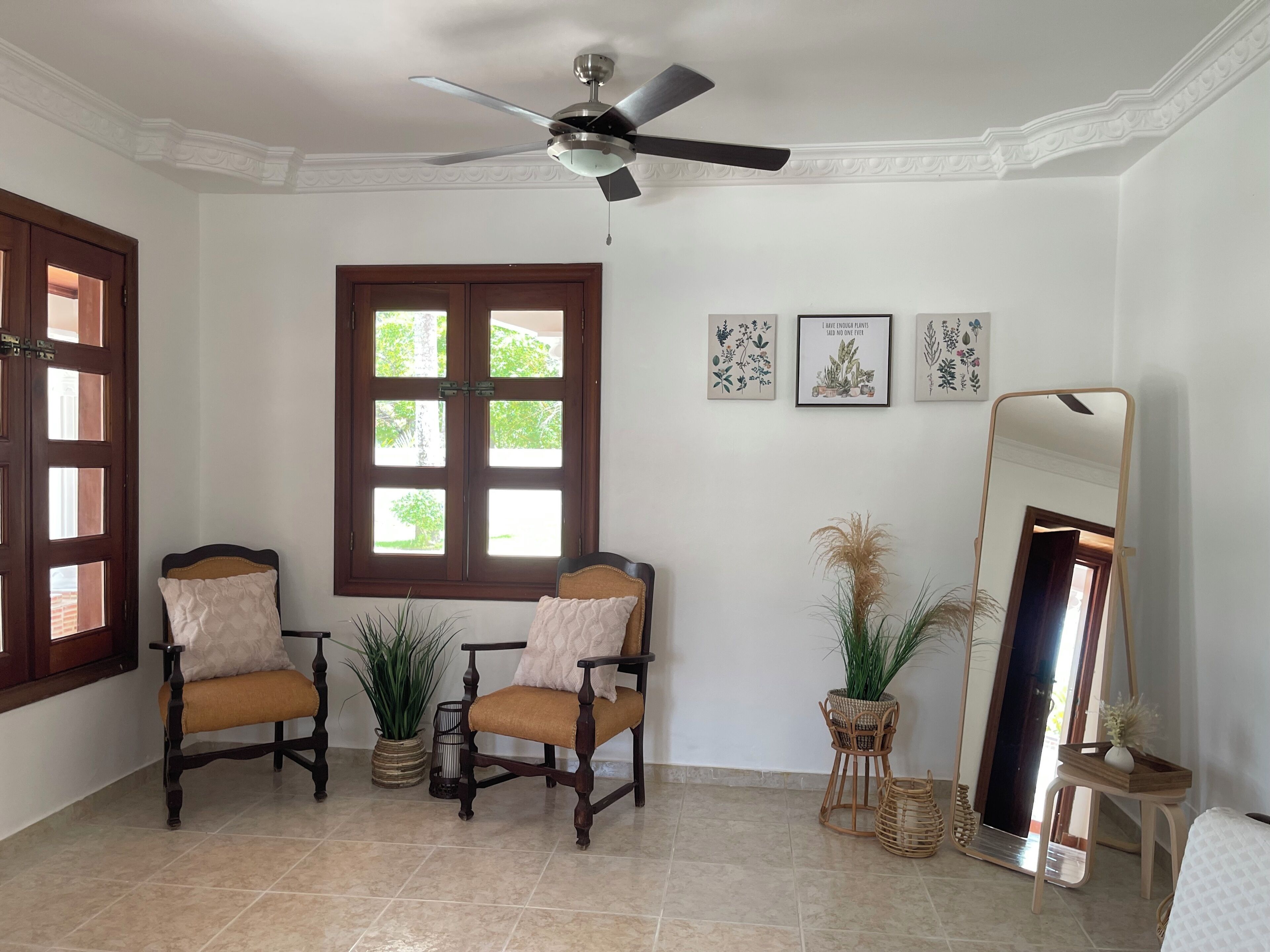 Una habitación con dos sillas de madera, un ventilador de techo, un ventanal grande y un espejo.