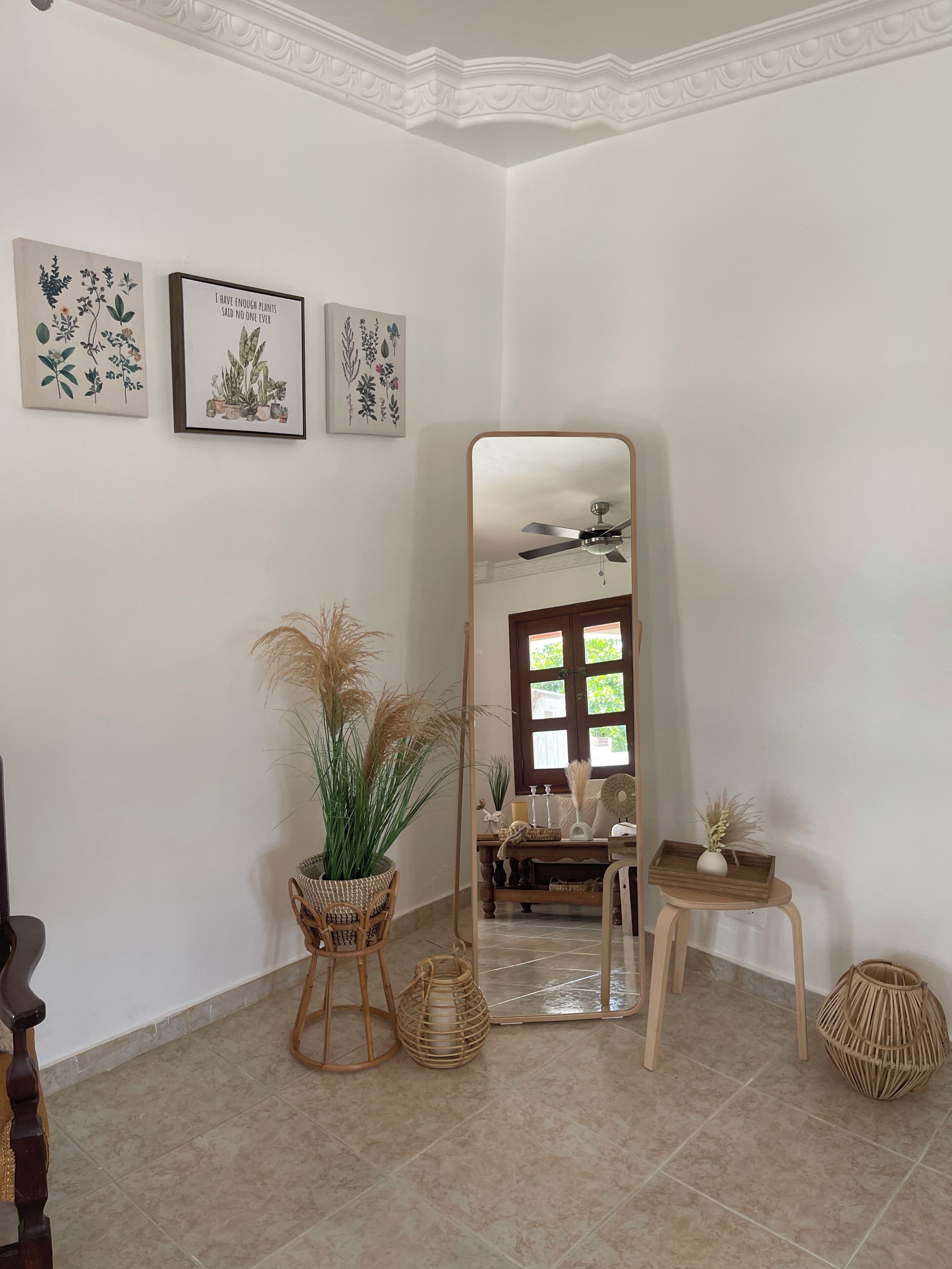 Een kamer met een grote spiegel, potplanten, rieten meubels en ingelijste botanische prints aan de muur.
