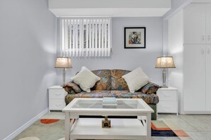 A modern living room with a sofa, two side tables, a glass coffee table, a framed picture, and a window with blinds. - Private 2BR Suite Near Ambassador Bridge (Windsor)