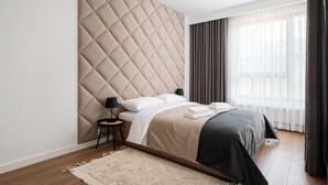 A modern bedroom with a tufted headboard, a bed with white linens, a bedside table with a lamp, and a large window with sheer curtains. - Wola Karolkowa Superior Apartment (Warszawa)