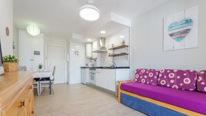 Een moderne keuken met witte kasten, een eethoek met een houten tafel en stoelen, en een zithoek met een paarse bank.