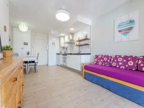 Une cuisine moderne avec des meubles blancs, un coin repas doté d’une table et de chaises en bois, et un salon avec un canapé violet.