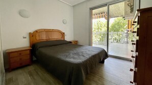 Quarto com cama, cabeceira de madeira, mesas de cabeceira, cômoda e porta de vidro corrediça para uma varanda.