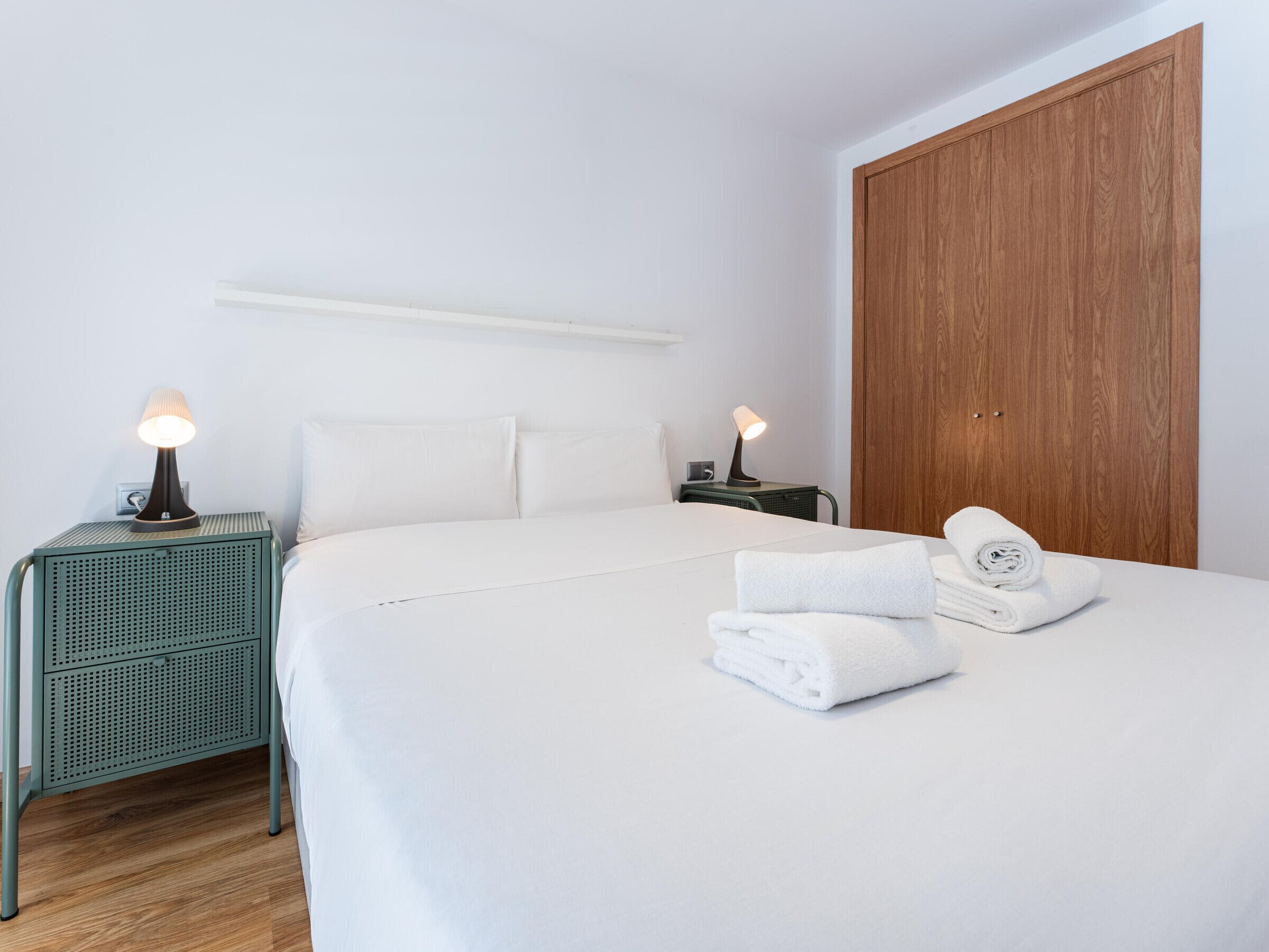 A neatly made bed with white linens, two bedside lamps, a wooden wardrobe, and a green metal nightstand with a lamp.