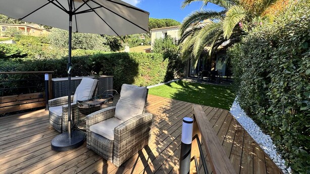 Eine Holzterrasse mit Korbmöbeln, einem großen Sonnenschirm und einem gepflegten Garten mit Hecke und Palme.
