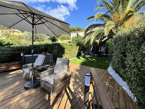 Um deck de madeira com mobiliário de vime, um grande guarda-sol e um jardim bem cuidado com um sebedeiro e palmeira.
