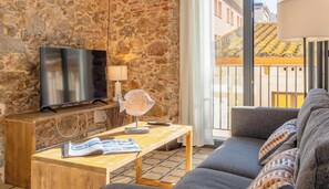 A living room with a stone wall, a flat-screen TV, a lamp, a fan, a sofa, and a view of buildings through a window. - Apartment with AC, central Palamós (Palamós)