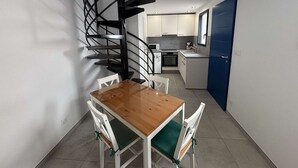 Eine kleine Küche mit weißen Schränken, einer Mikrowelle und einem Spülbecken. Ein Holztisch wird von weißen Stühlen begleitet. Im Hintergrund ist eine schwarze Wendeltreppe zu sehen.