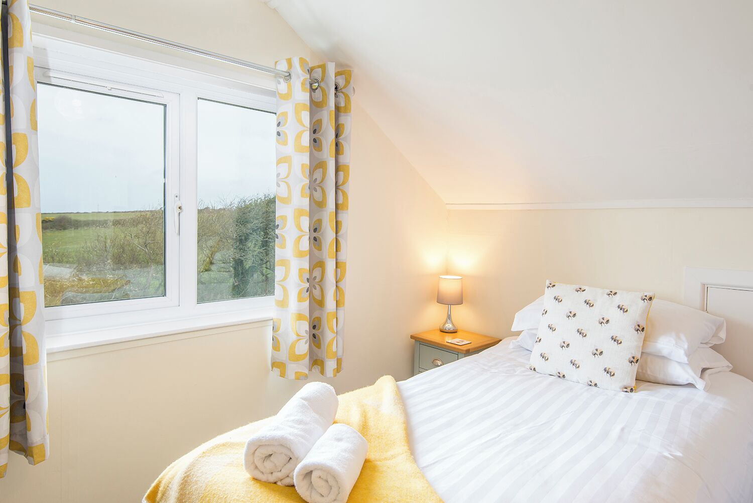 A bedroom with a bed, pillows, a nightstand, a lamp, and a window with curtains.