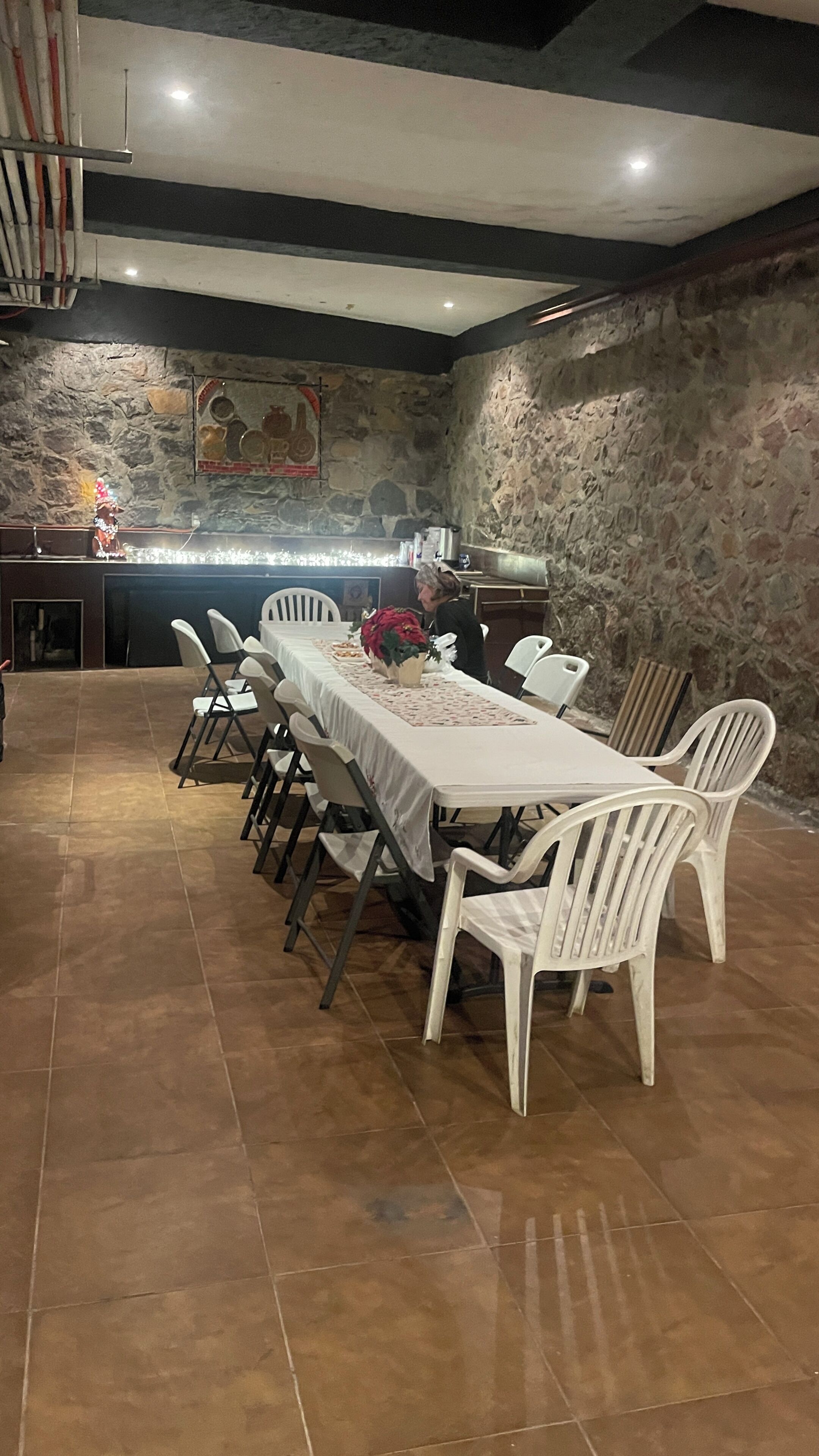 Un espace repas avec une longue table, des chaises blanches, un mur en pierre et un sol carrelé.