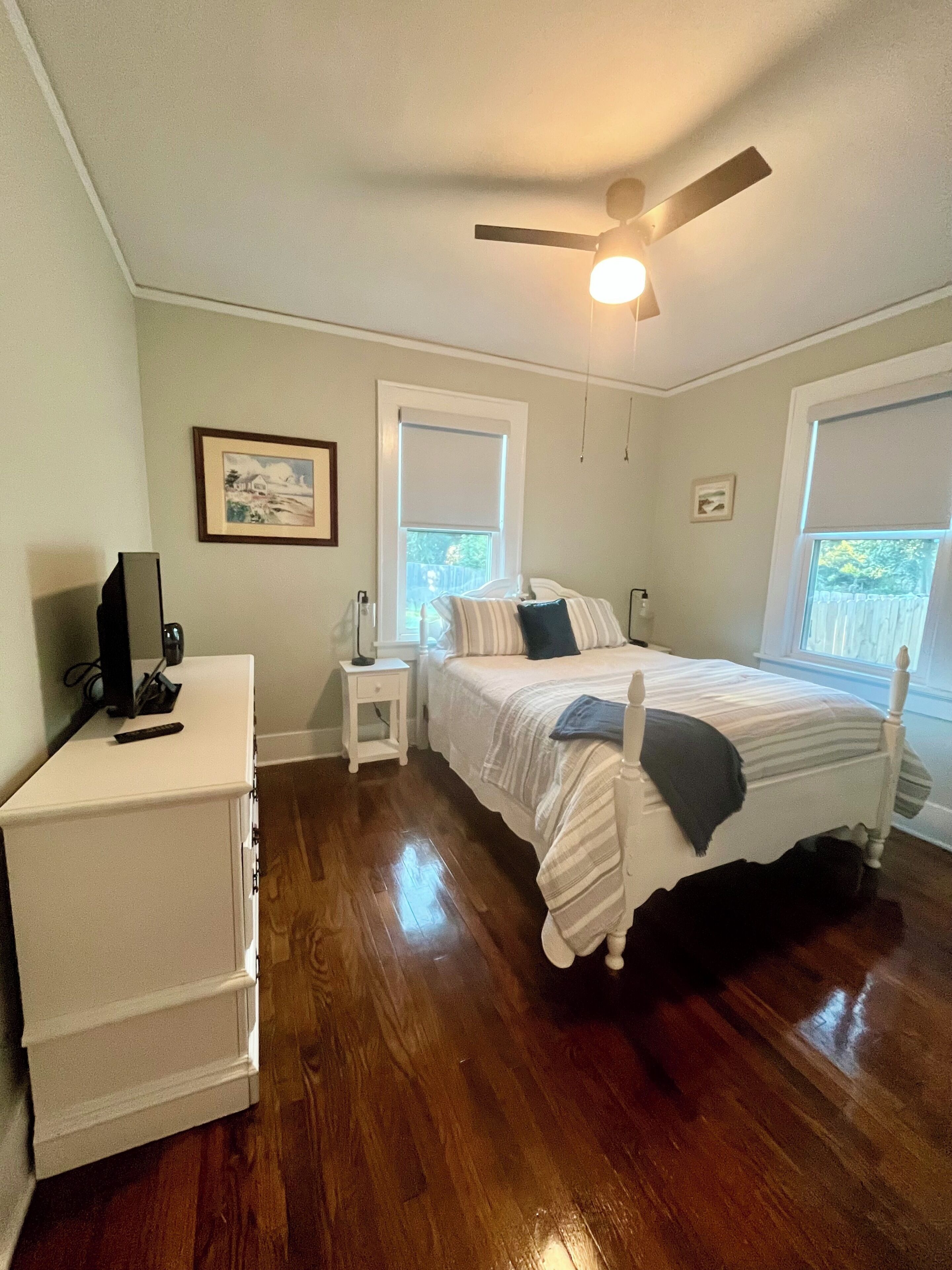 Une chambre à coucher comprenant un lit, un ventilateur de plafond, une table de chevet, une commode et une fenêtre.