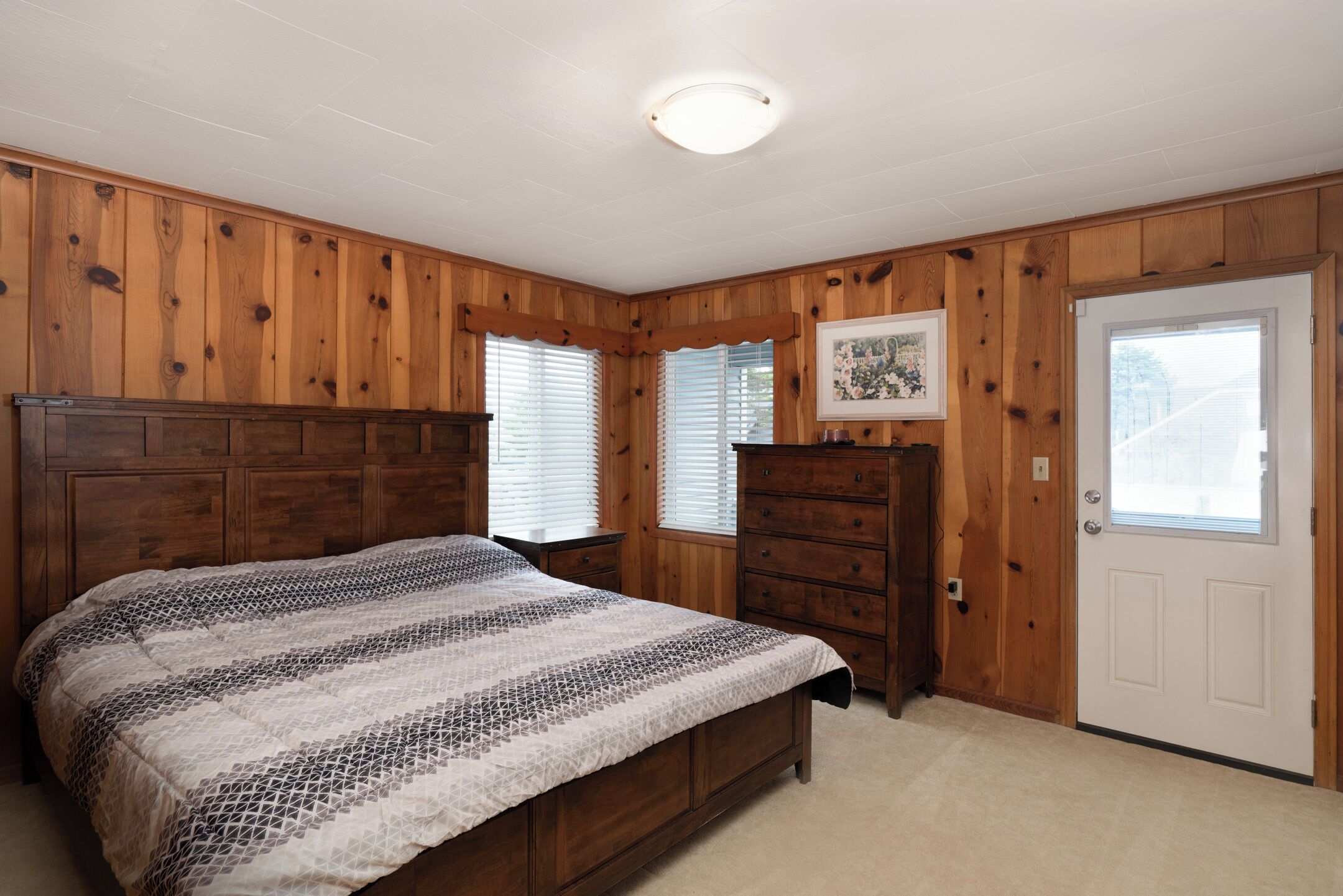 A bedroom with a wooden paneled wall, a bed with a patterned bedspread, a dresser, and a door with a window.