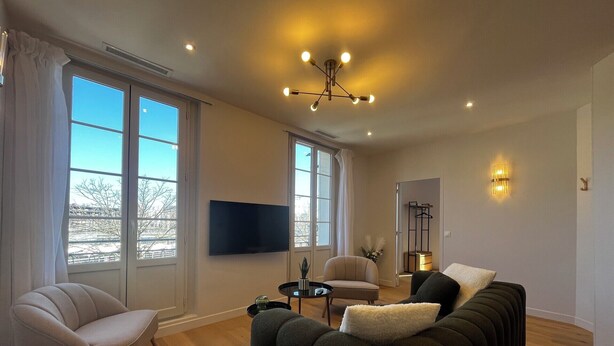 Een woonkamer met een bank, fauteuil, televisie en grote ramen.