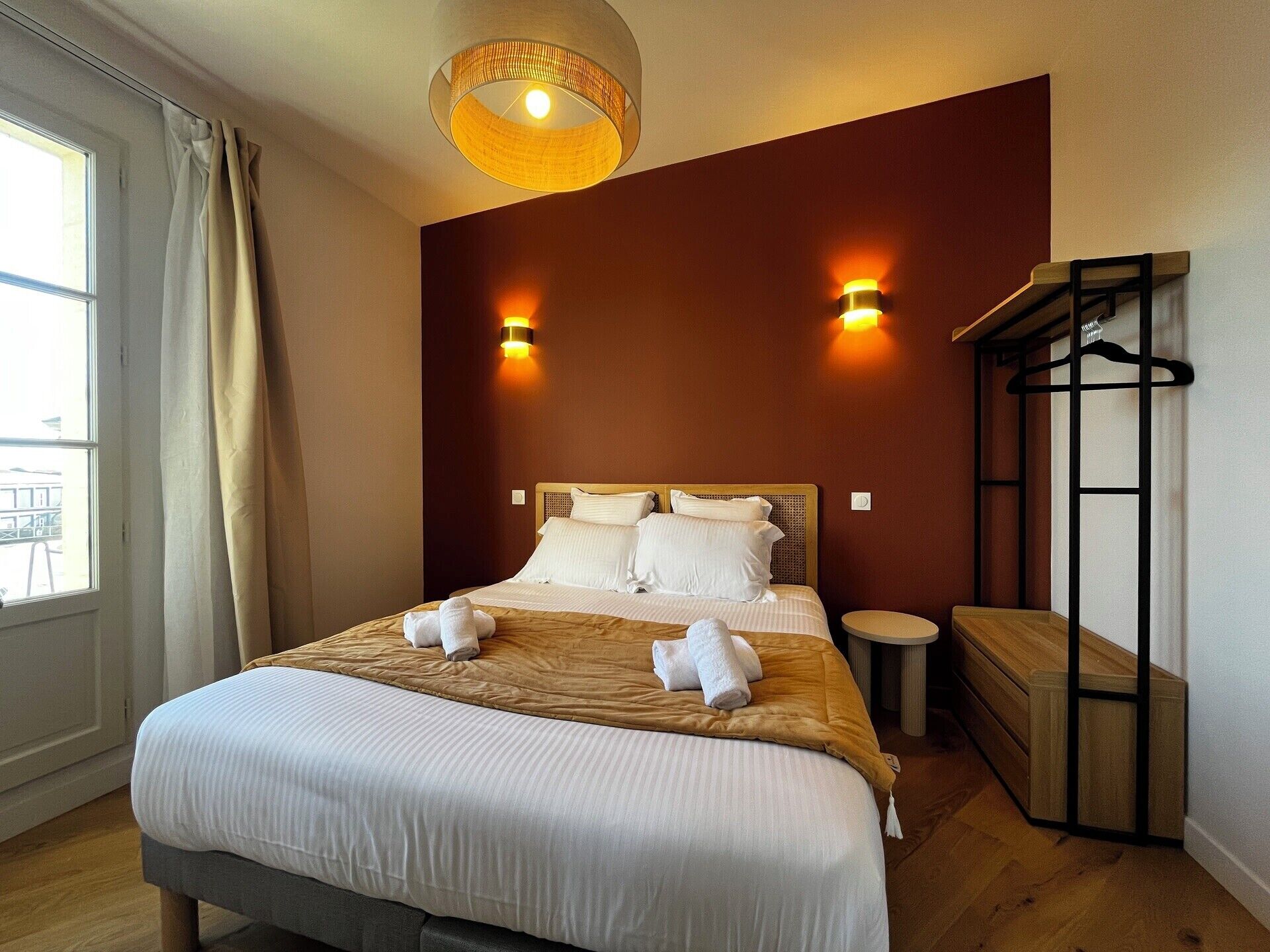 Ein Hotelzimmer mit einem Bett, zwei Wandlampen, einer runden Deckenleuchte, einer hölzernen Bank und einem Kleiderständer.