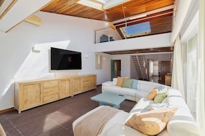Amplia sala de estar con un televisor de pantalla plana grande, armarios de madera, un sofá blanco y una escalera que conduce a un nivel superior.
