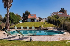 Una piscina con sillas de descanso, un patio de piedra y una casa con tejado de tejas al fondo.