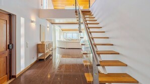 Un interior moderno con una escalera de madera, barandilla de vidrio y vista a una sala de estar.