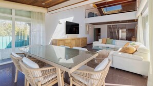 Un salón amplio con una mesa de vidrio, sillas de mimbre, un sofá blanco y un televisor montado en la pared.