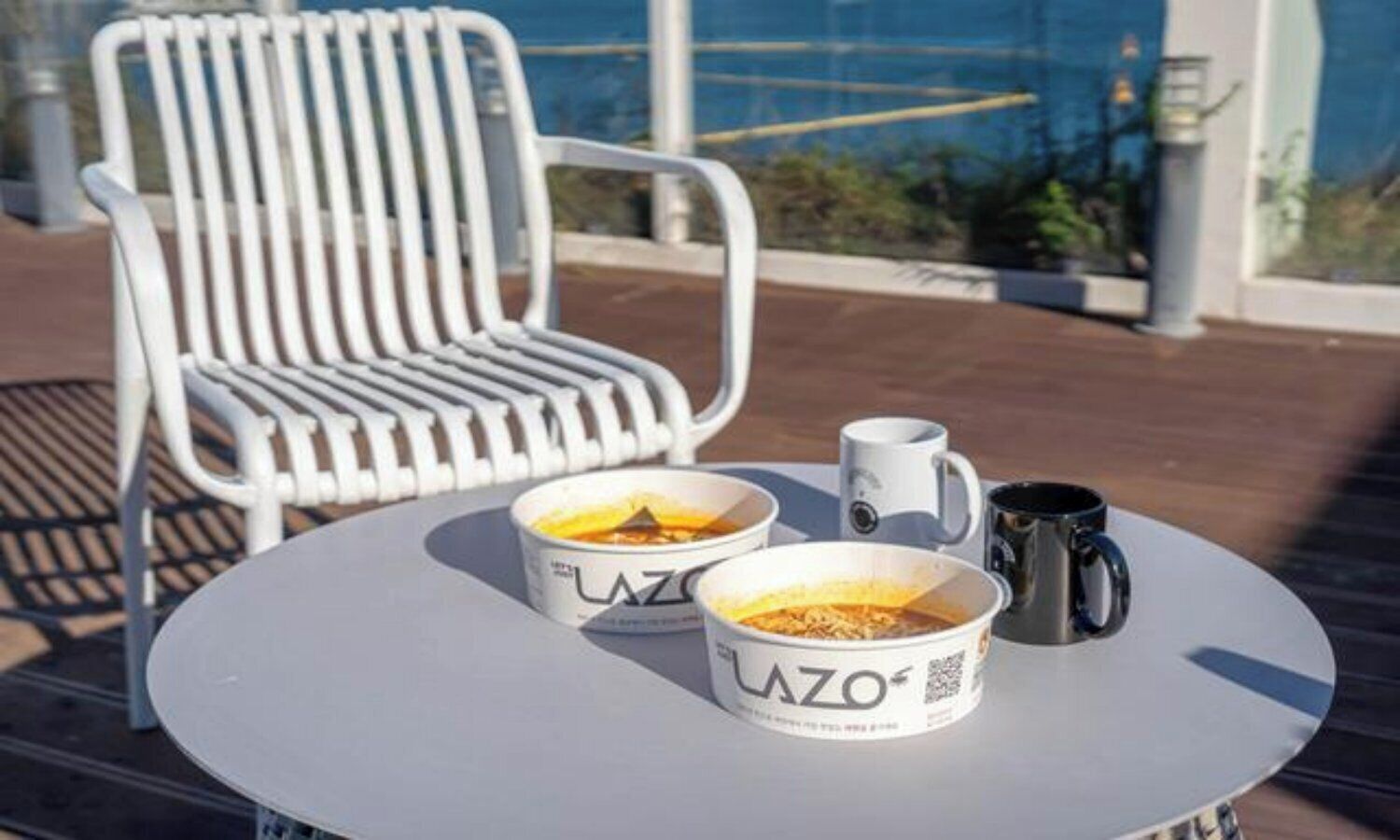 Une table avec deux tasses à café et une tasse noire, une chaise blanche et une vue sur un bâtiment et de la verdure.