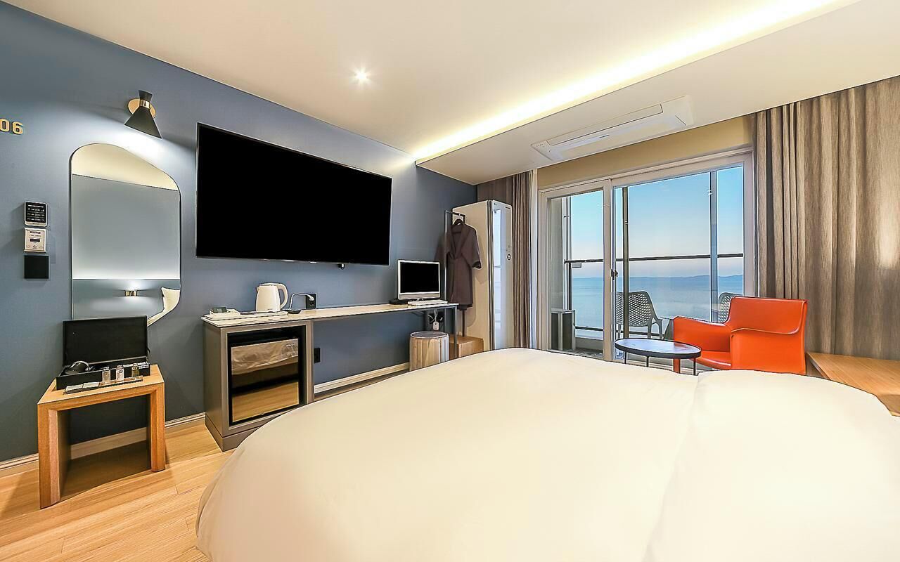 A modern hotel room with a large bed, a flat-screen TV, a small table, a chair, and a balcony with a view.