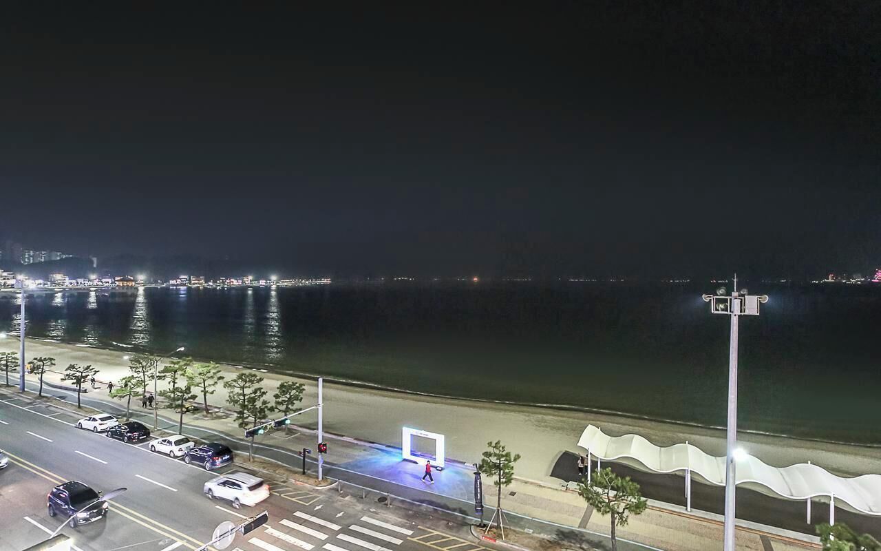 A nighttime view of a waterfront area with a road, parked cars, and a pedestrian crossing.