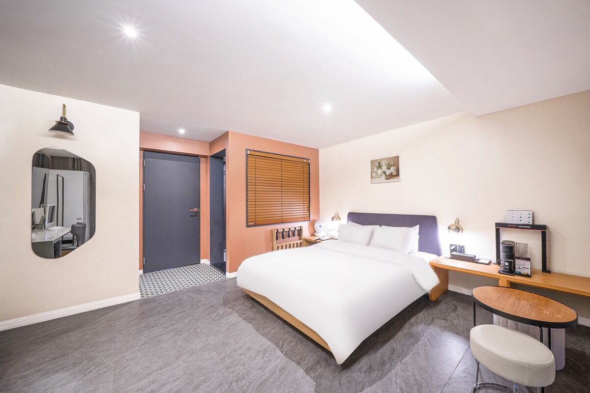 A hotel room with a bed, bedside table, chair, and a desk with a computer.