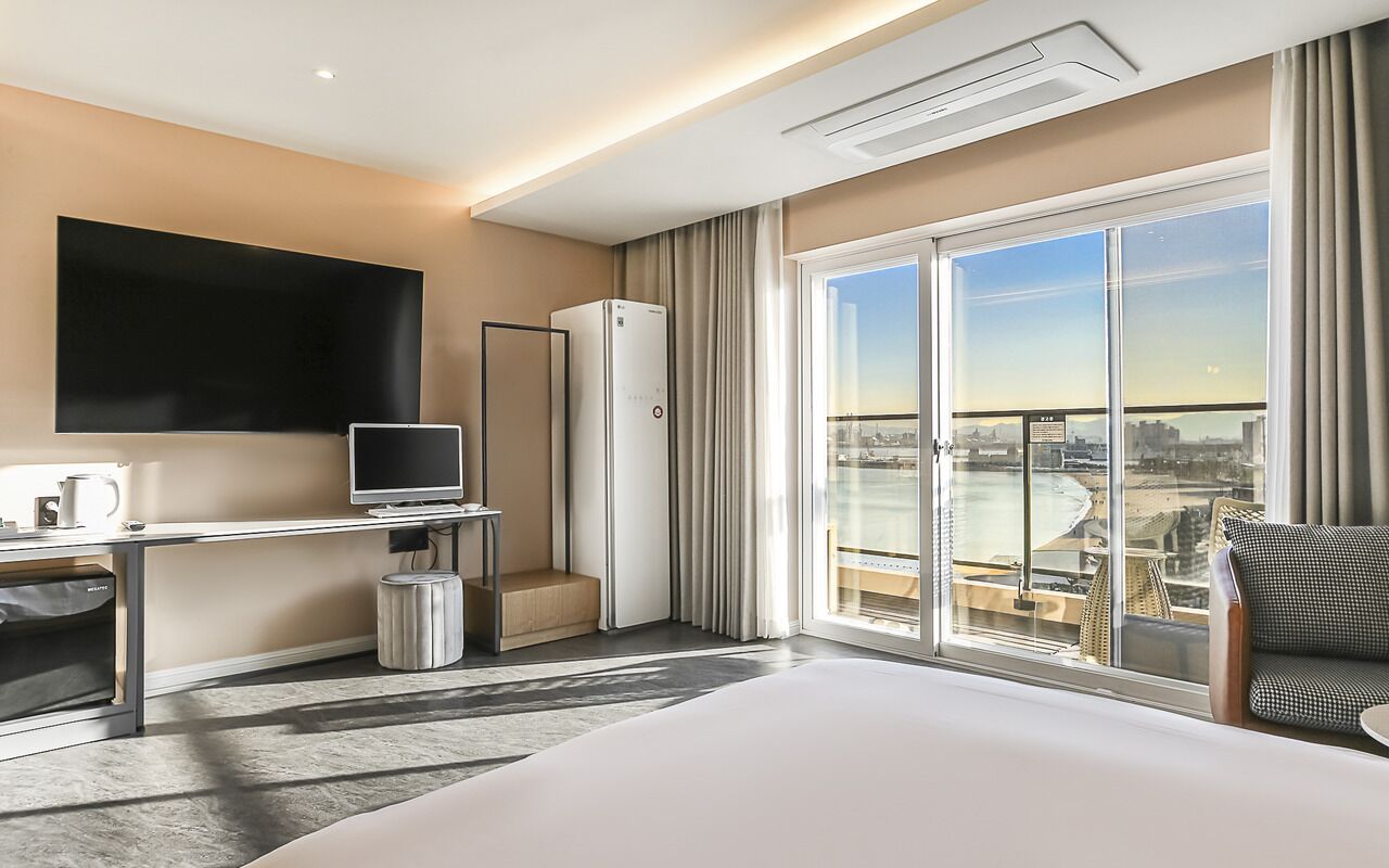 Une chambre d’hôtel dotée d’un grand lit, d’un téléviseur à écran plat, d’un bureau avec un ordinateur, d’une poubelle et d’une porte-fenêtre coulissante donnant sur un balcon avec vue sur la ville.