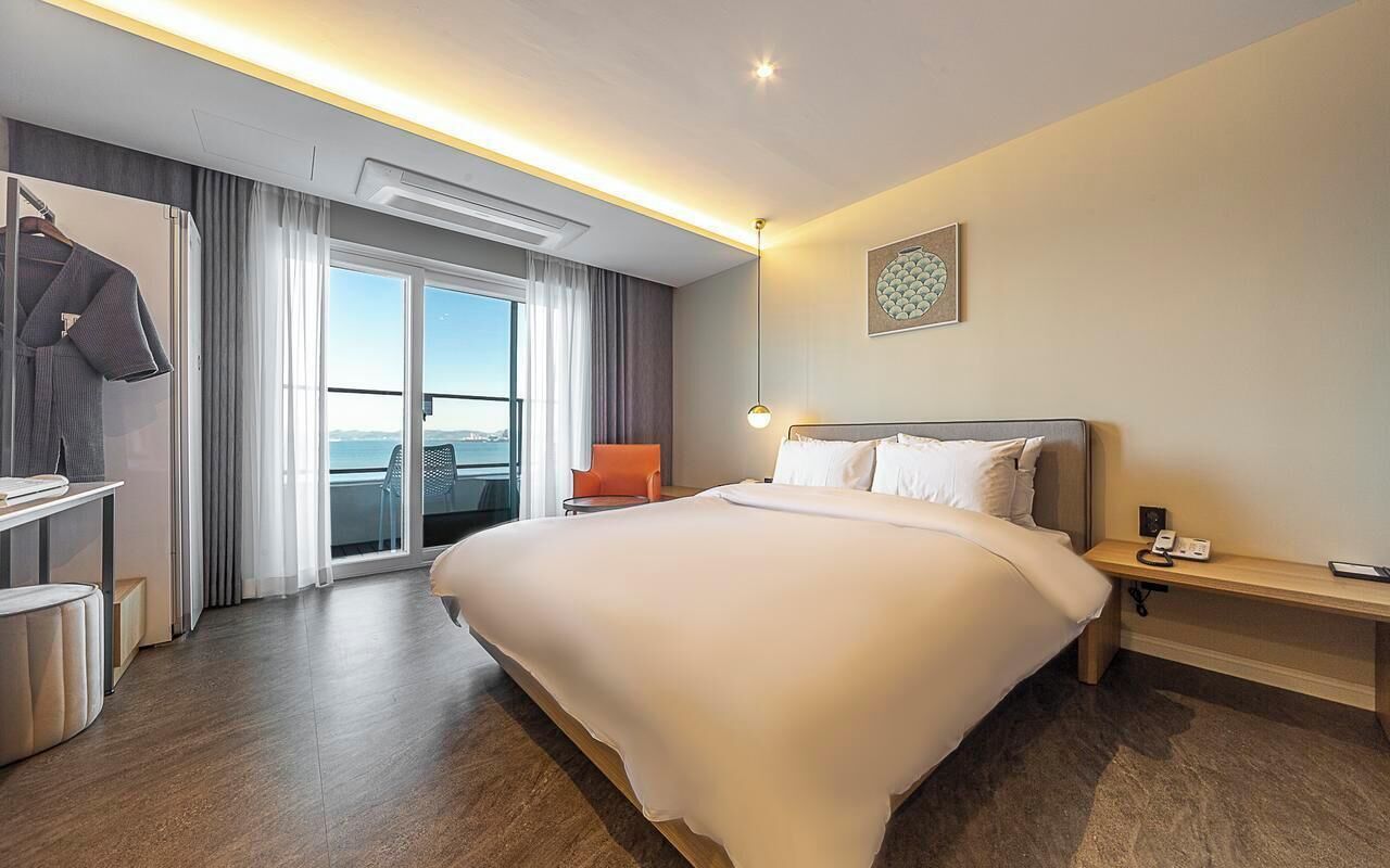 A hotel room with a large bed, a desk, a chair, a wardrobe, and a view of the sea.