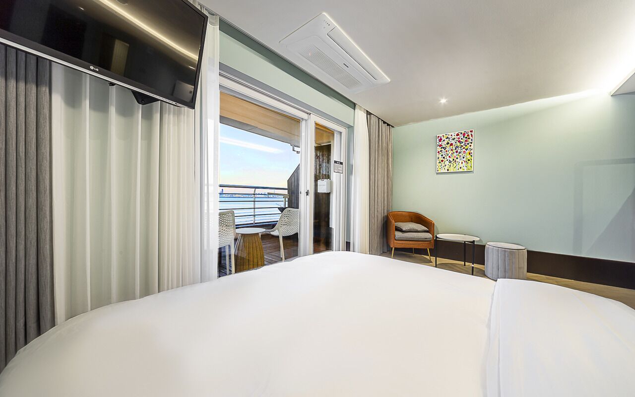 Habitación de hotel con cama, una silla, una mesita, un cuadro y una puerta corrediza que da a un balcón.