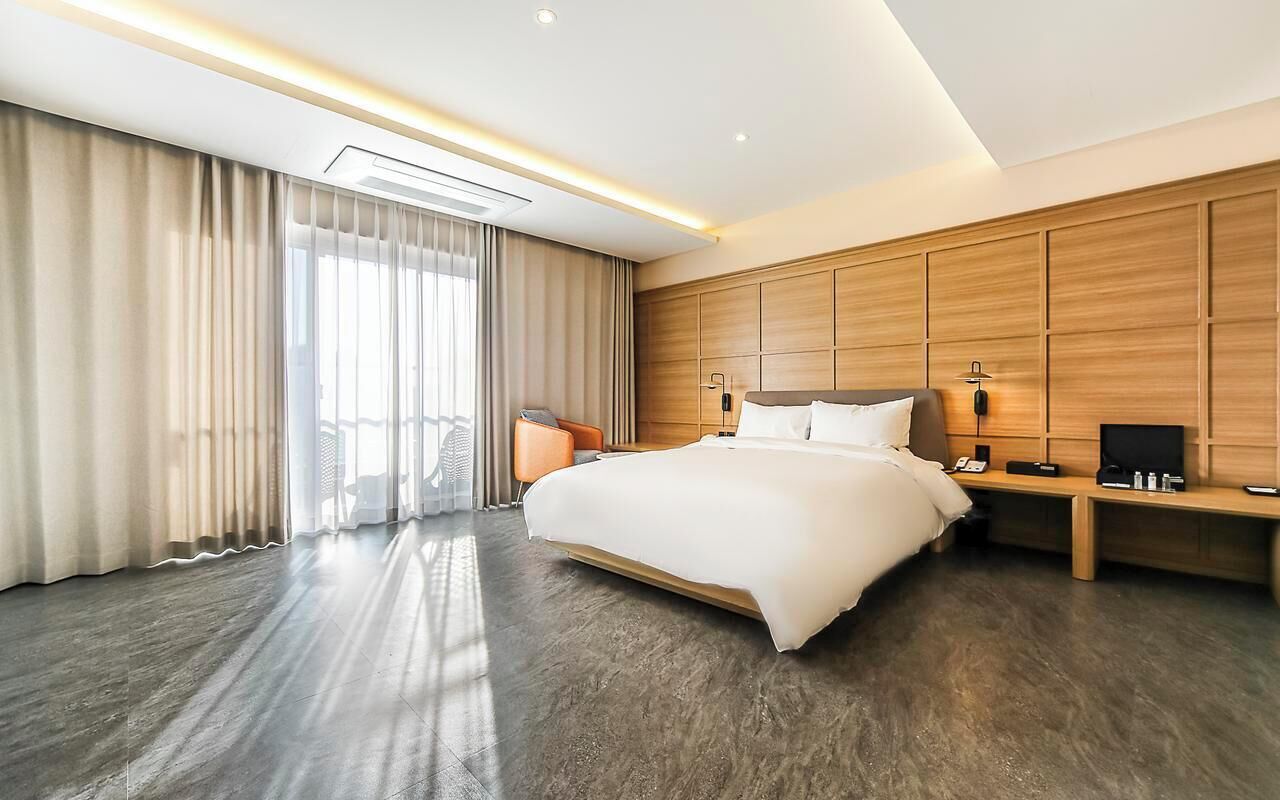 A modern hotel room with a large bed, a wooden headboard, a desk with a chair, and a large window with curtains.