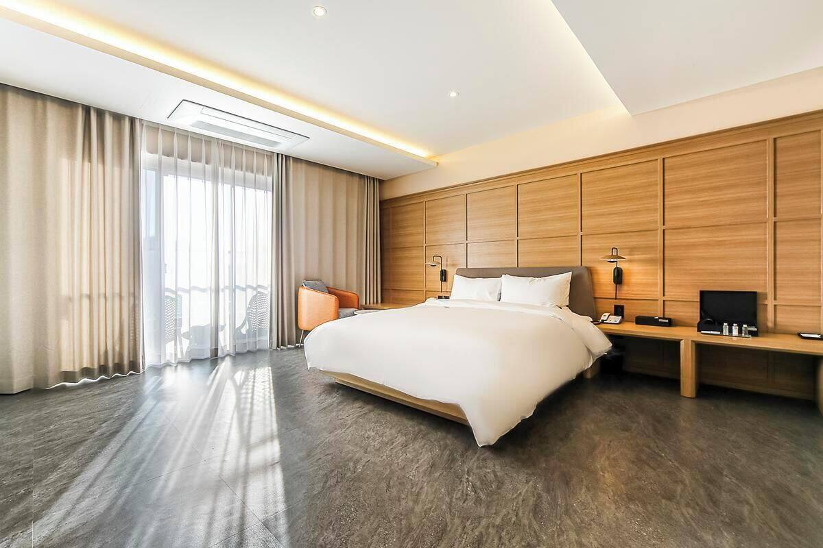 A modern hotel room with a large bed, a wooden headboard, a desk with a chair, and a large window with curtains.