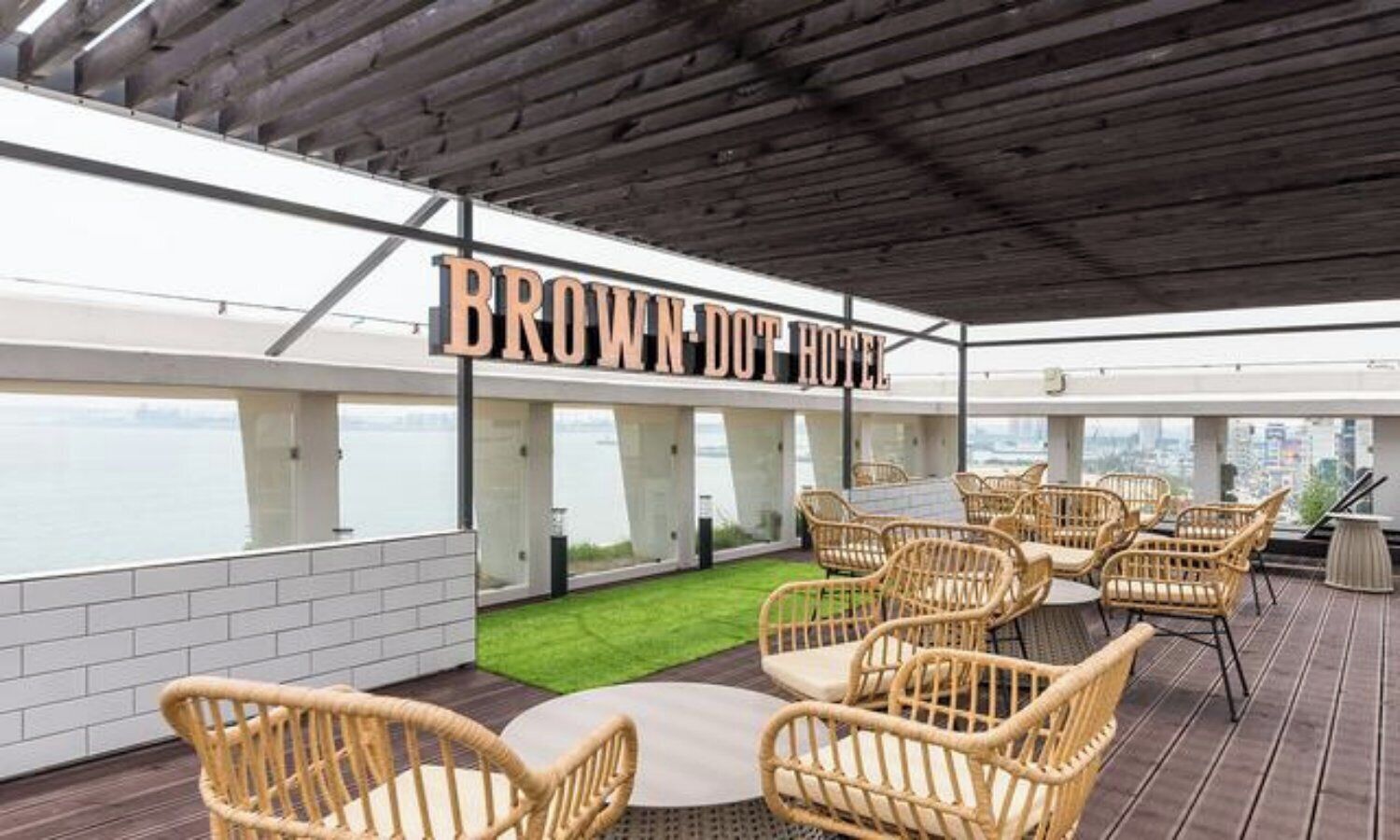 A rooftop terrace with wicker furniture, a sign reading 'BROWN DOT HOTEL,' and a view of the cityscape.