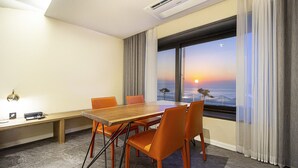 A hotel room with a wooden table, orange chairs, a bench, a desk with a lamp, and a large window with a view of the sunset over the ocean.