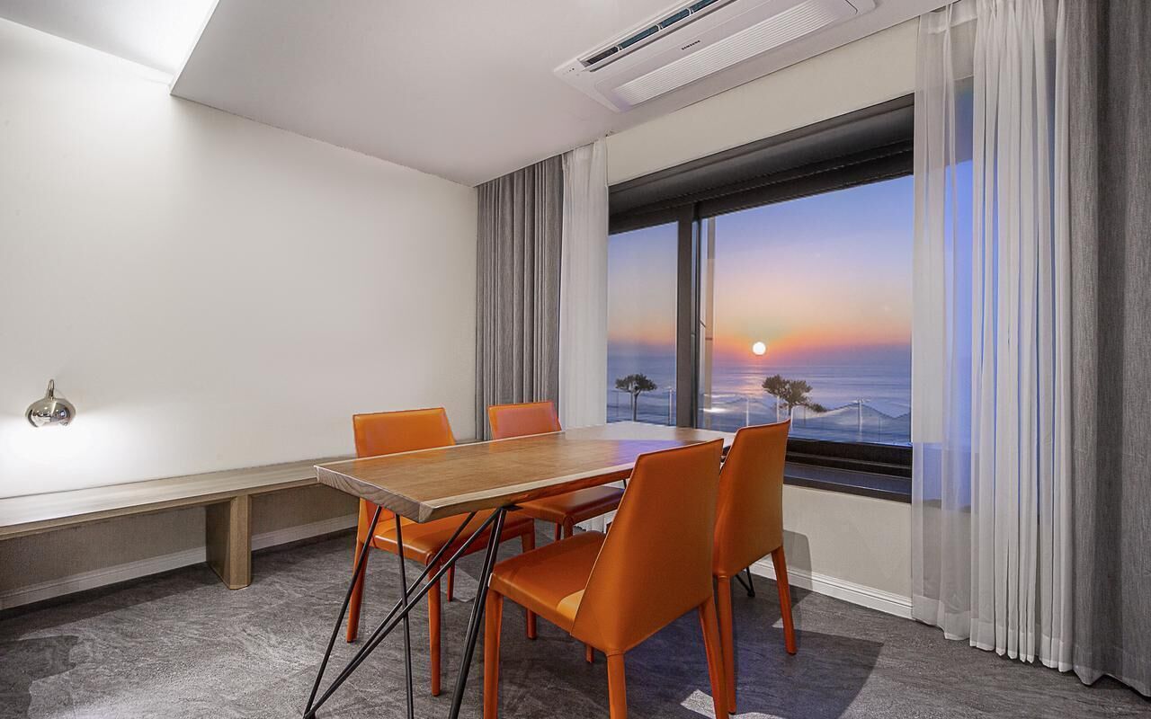 A conference room with a wooden table, orange chairs, a bench, and a large window with a sunset view.