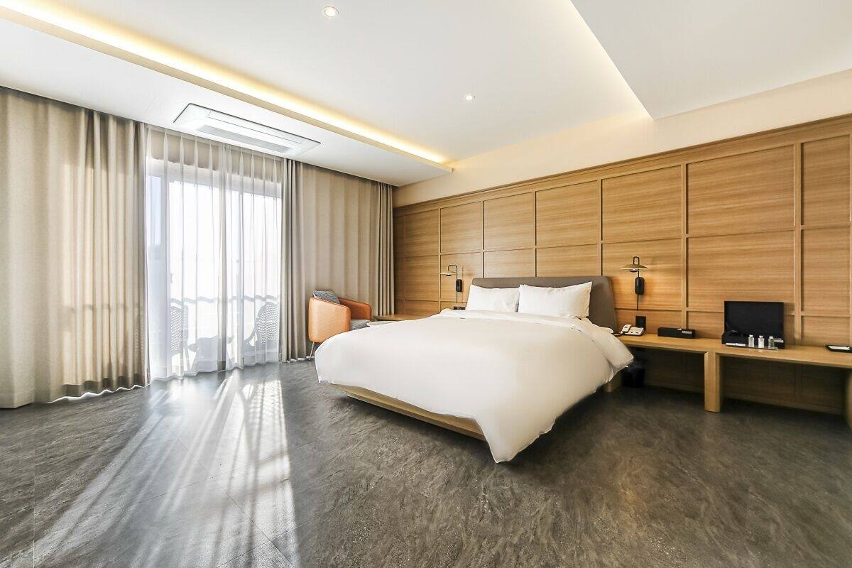 A modern hotel room with a large bed, a wooden headboard, a bedside table with a lamp, and a large window with curtains.