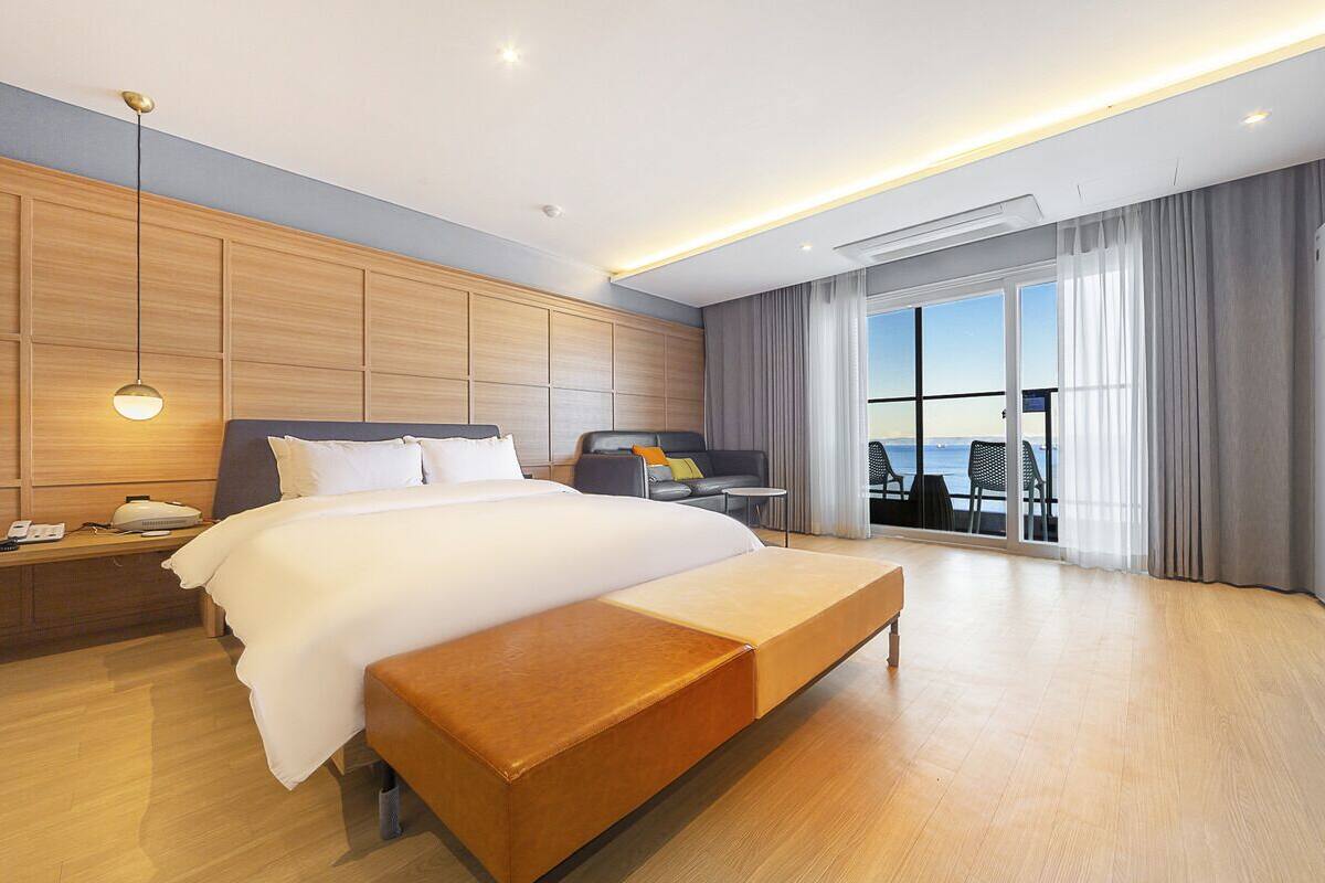A modern hotel room with a large bed, a bedside table, a bench, a desk, and a view of the sea through large windows.