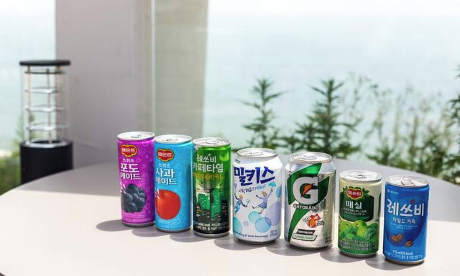 A variety of beverage cans on a table with a view of trees and a building in the background.