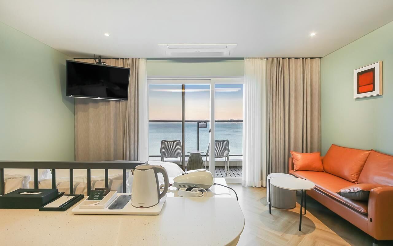 Een hotelkamer met balkon, flatscreen televisie, bank, tafel met waterkoker, en uitzicht op zee.