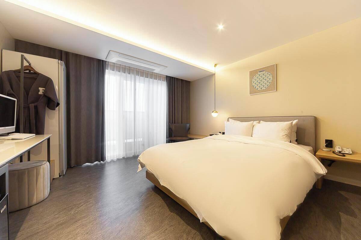 A hotel room with a large bed, a desk with a chair, a TV, a wardrobe, and a bedside table with a lamp.