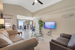 A spacious living room with a sofa, armchairs, a glass coffee table, a TV mounted on the wall, and a potted plant. - Sunny Stay Near St. Pete Beaches (Pinellas Park)