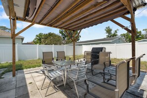 Een overdekt terras met een barbecue, metalen stoelen en een glazen tafel.